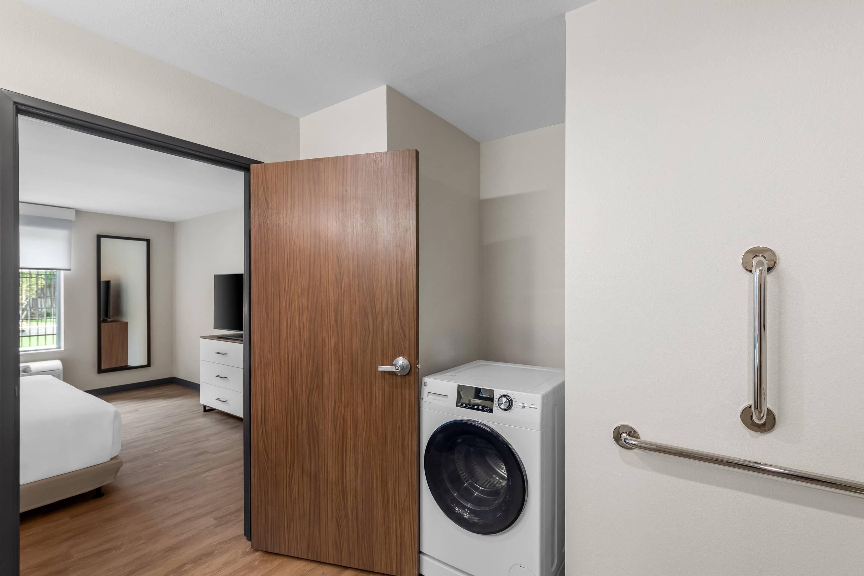 Una habitación de hotel moderna con una puerta de madera, lavadora, pared blanca y barandilla de metal.