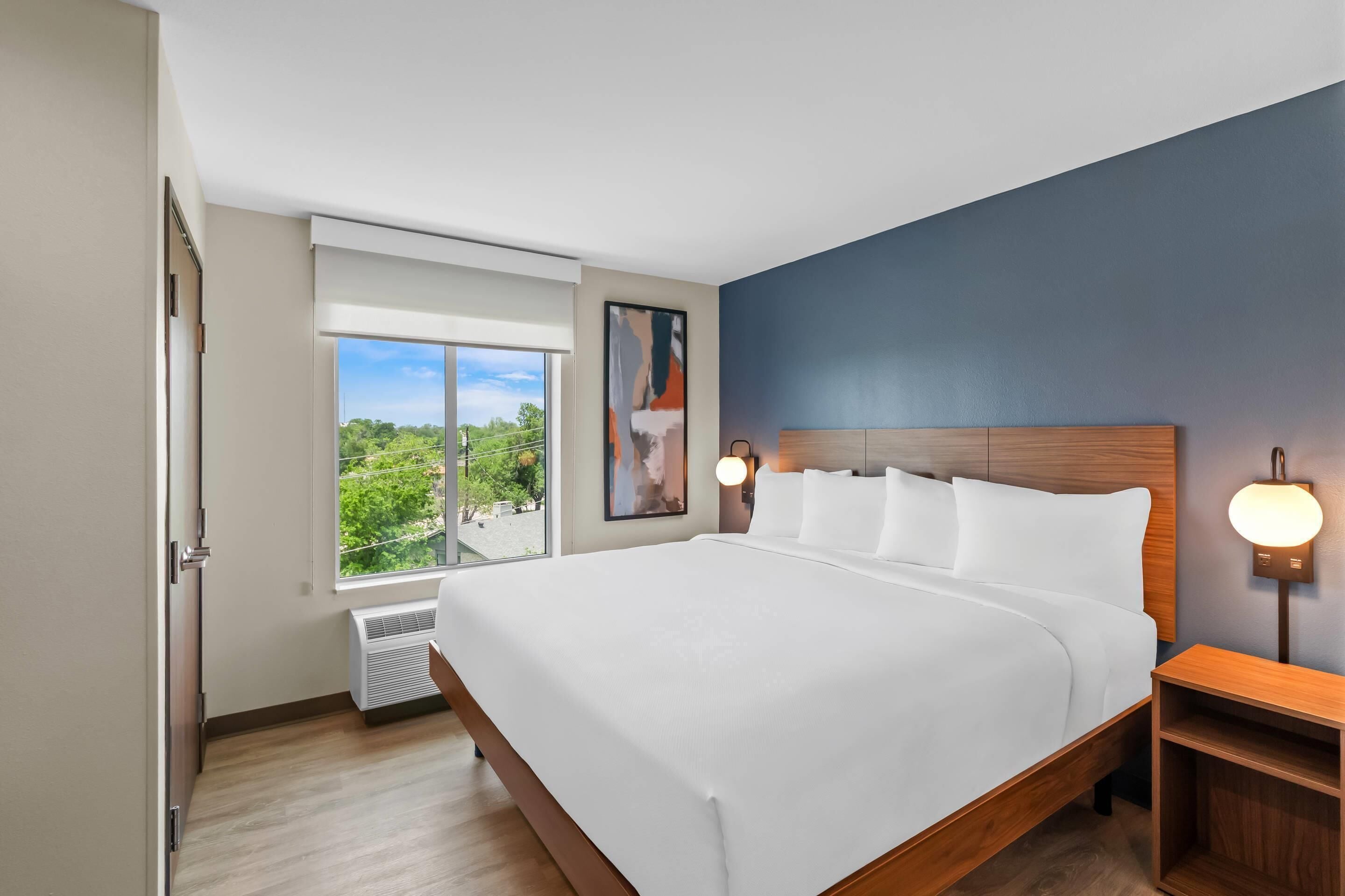 Une chambre d’hôtel moderne avec un grand lit, une table de chevet, une lampe fixée au mur et une fenêtre donnant sur un paysage verdoyant.