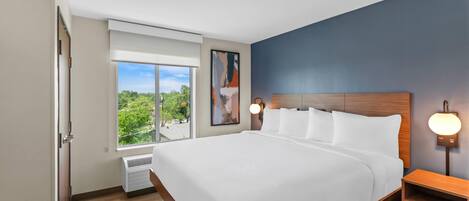 Une chambre d’hôtel moderne avec un grand lit, une table de chevet, une lampe fixée au mur et une fenêtre donnant sur un paysage verdoyant.