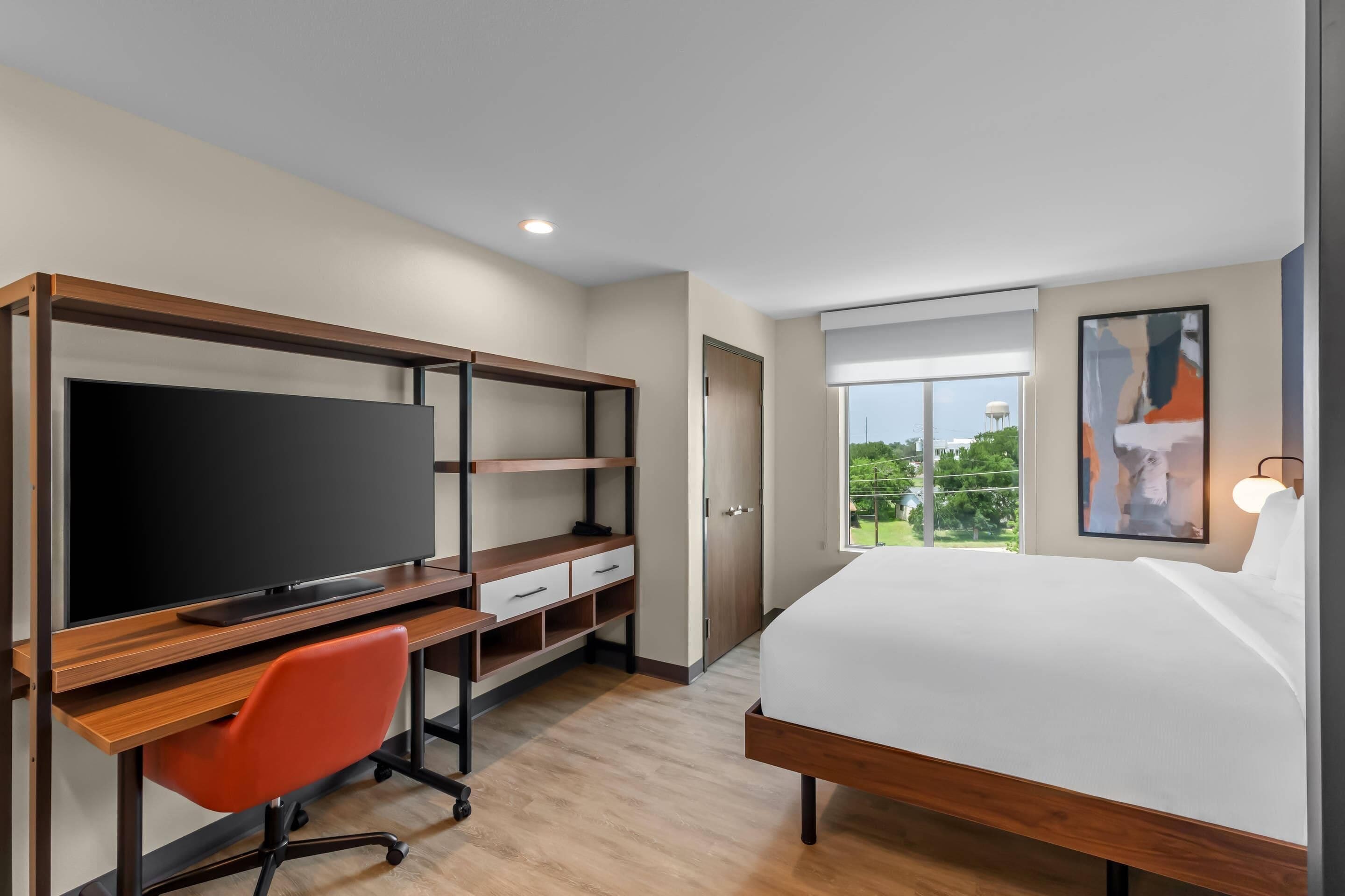 Una habitación de hotel moderna con una cama grande, un escritorio con televisión de pantalla plana, una silla de oficina roja y una ventana con vistas a zonas verdes y una torre de agua.
