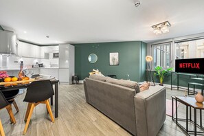 Un salon moderne avec un canapé gris, un coin repas avec une table et des chaises en bois, une cuisine en arrière-plan et une grande fenêtre offrant une belle vue.