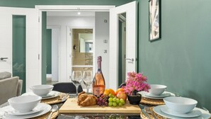 Ein Esstisch, gedeckt mit weißen Schüsseln, Tellern, Besteck, einer Rosé-Weinflasche, Früchten und einem Croissant.