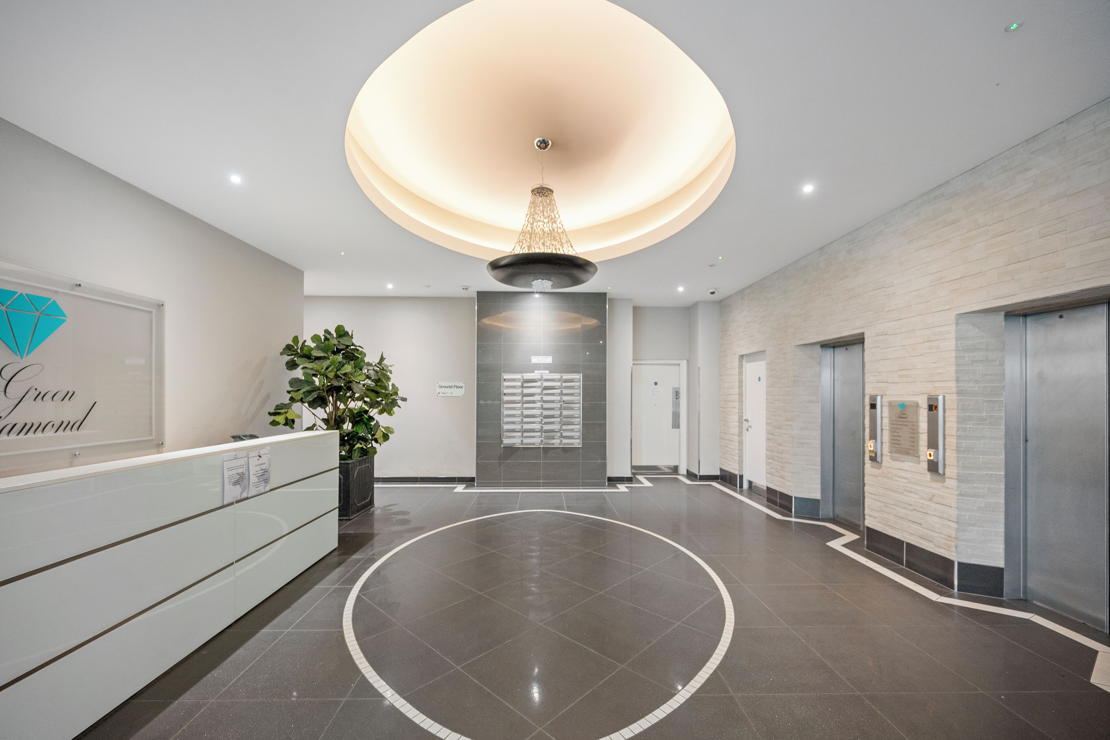 A modern reception area with a central circular floor design, a reception desk, a large chandelier, and a wall-mounted information board.