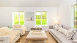 Una sala de estar moderna con un sofá, sillones y una mesa de centro. Hay dos ventanas con vistas a un jardín verde.