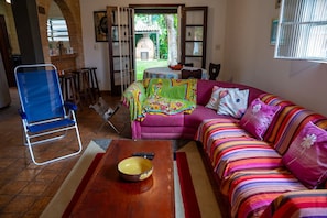 Un salon comprenant un canapé, un fauteuil bleu, une table basse en bois et donnant sur un espace extérieur aménagé avec une table et des chaises.
