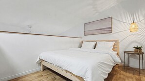 A bedroom with a wooden bed, white bedding, a small table with a plant, a hanging light fixture, and a painting on the wall. - Loft le Mercier by Gestion ELITE (Mont-Tremblant)