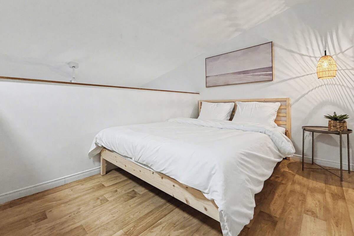 A bedroom with a wooden bed, white bedding, a small table with a plant, a hanging light fixture, and a painting on the wall.