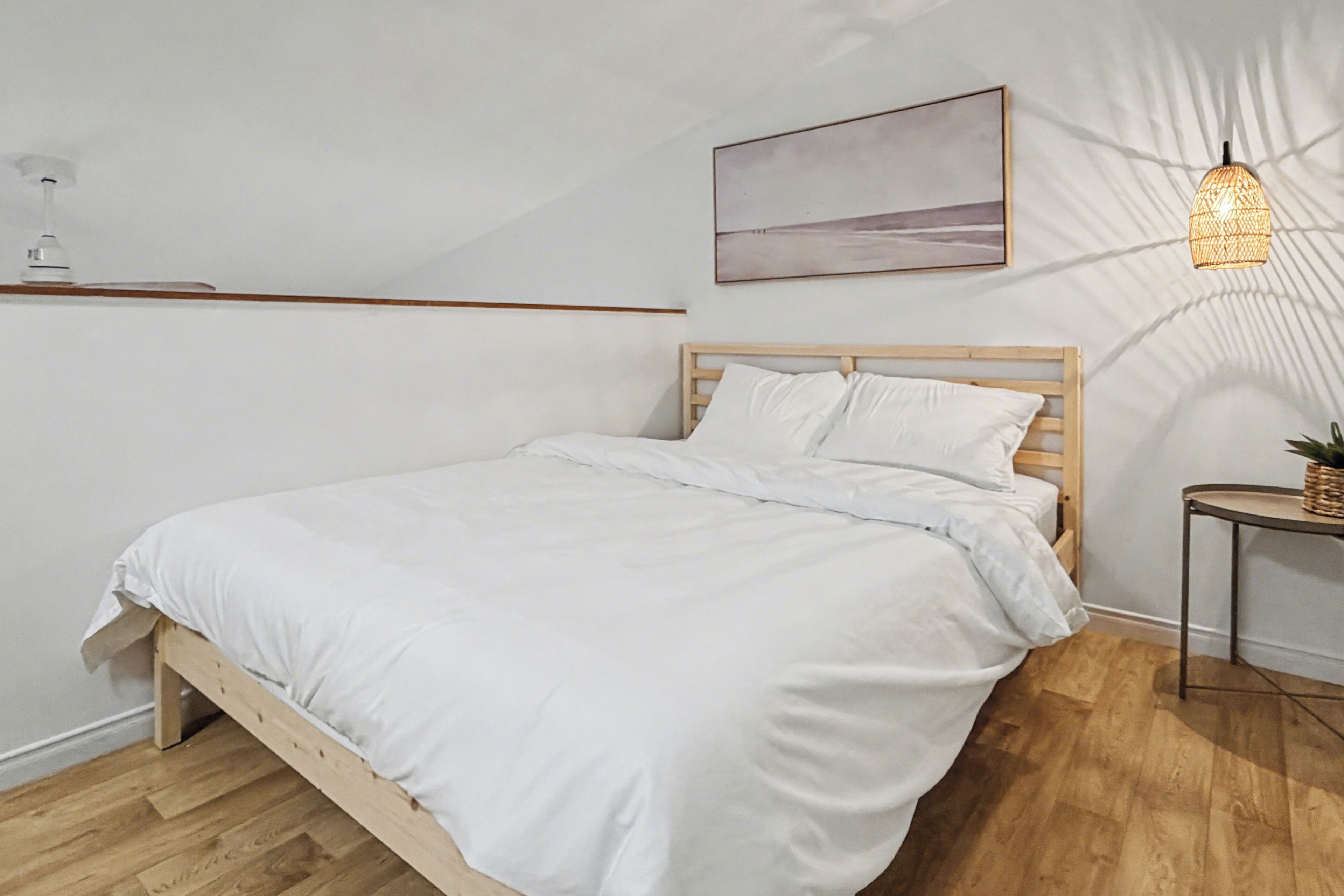 Un dormitorio con una cama de madera, ropa de cama blanca, un ventilador de techo, una lámpara de pared y un cuadro sobre la cama.