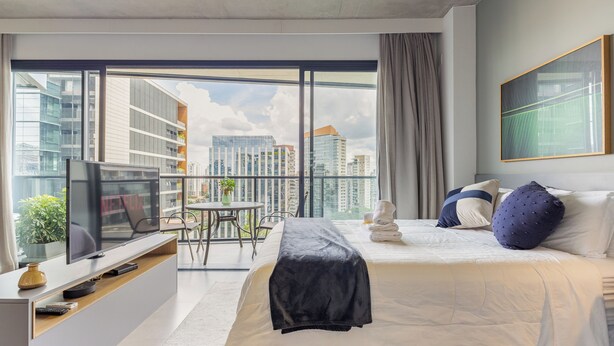 A modern hotel room with a large bed, a TV on a wooden console, a balcony with a view of the city, and a dining area with a table and chairs. - Luxo na Faria Lima - Millenium (São Paulo)