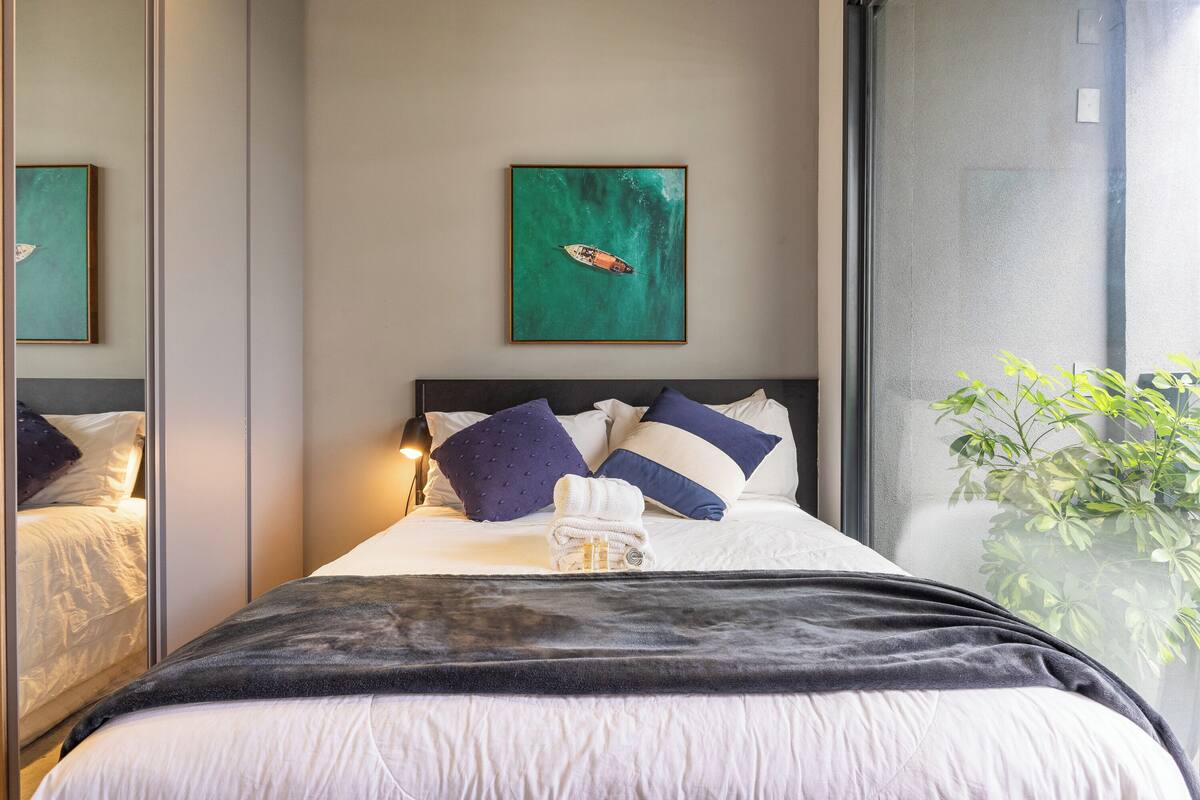 Een netjes opgemaakt bed met een donkere plaid, een ingelijste afbeelding erboven en een potplant aan de rechterkant.
