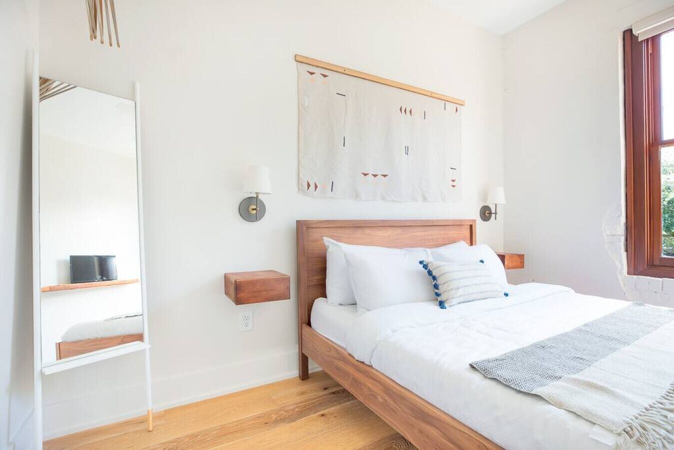 Un dormitorio con cama de madera, ropa de cama blanca, una obra de arte en la pared, lámparas de noche y una ventana con vistas.