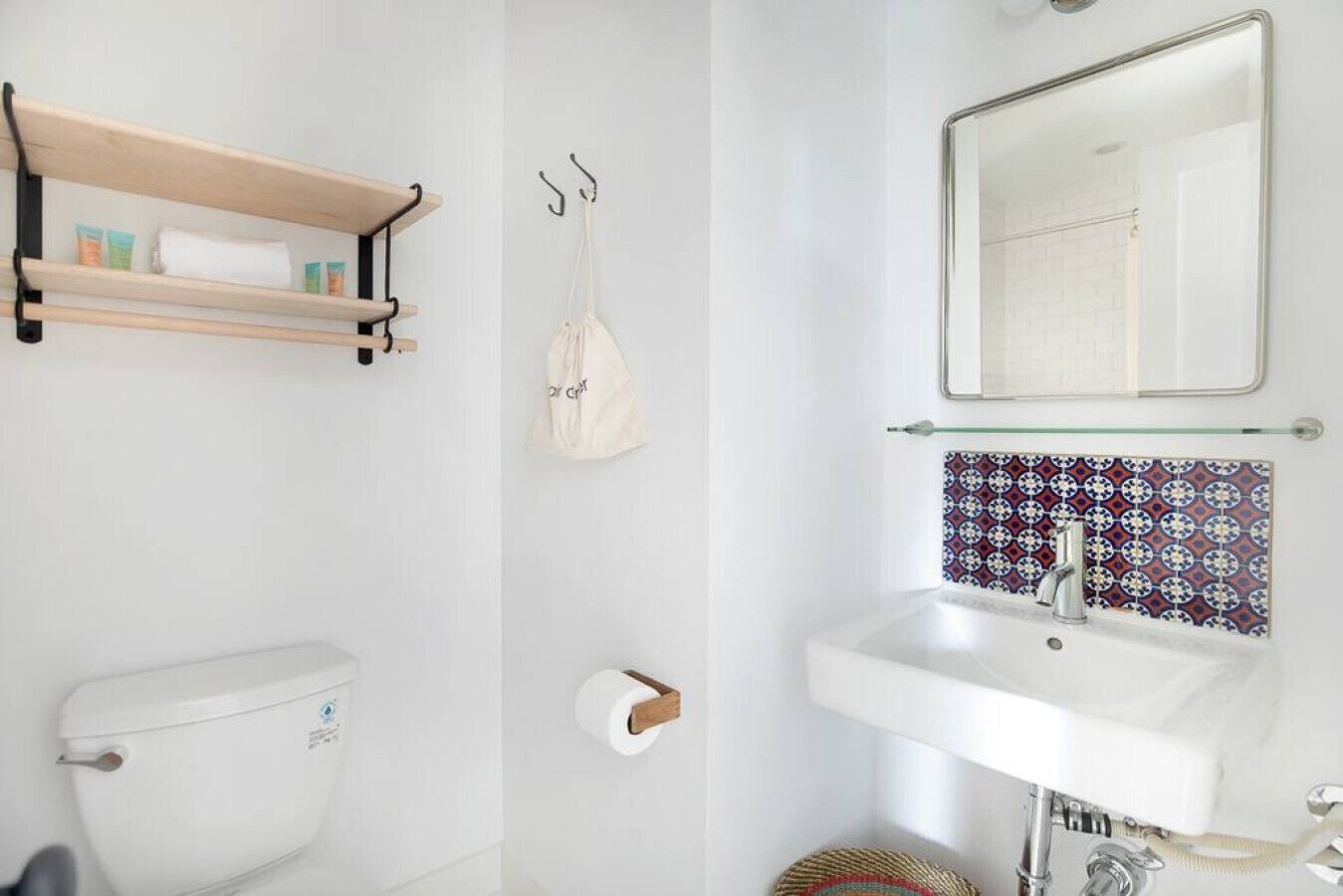 A bathroom with a toilet, a sink, a mirror, a shelf with toiletries, and a hanging bag.