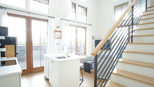A modern kitchen with a central island, a built-in sink, and a staircase with wooden steps and metal railings. - The Windrose (Savannah)