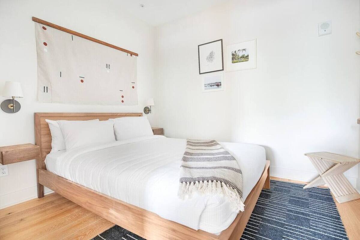 Une chambre à coucher bien rangée, avec un lit en bois, du linge de lit blanc, un plaid à rayures et une petite table de chevet surmontée d’une lampe.