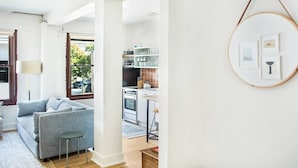 A modern living room with a grey sofa, a small table, and a kitchen area with white cabinets and a stove. - The Windrose (Savannah)