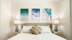 A neatly made bed with a quilted white bedspread, two bedside tables with lamps, and three framed beach art pieces on the wall. - #304 Resort 3BR/2BA Pool/Spa WindsorHill Disney (Kissimmee)