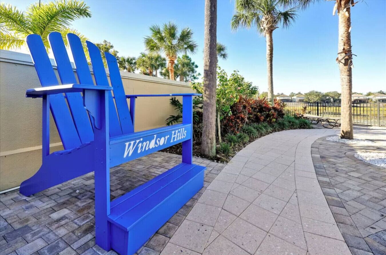 A blue Adirondack chair with the text "Windsor Hills" on a paved pathway surrounded by palm trees and greenery.