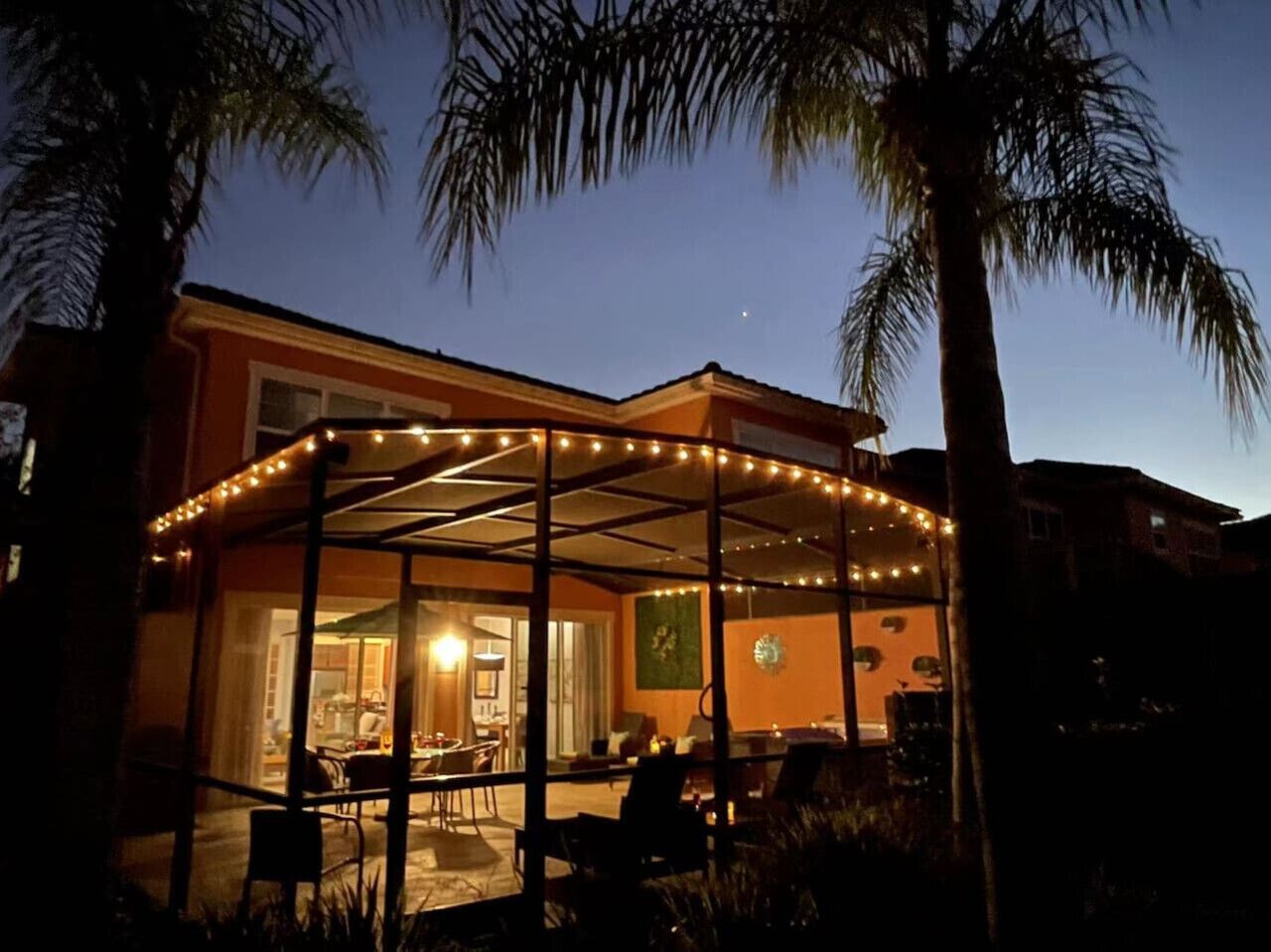 A covered outdoor dining area with string lights, palm trees, and a view of the interior.