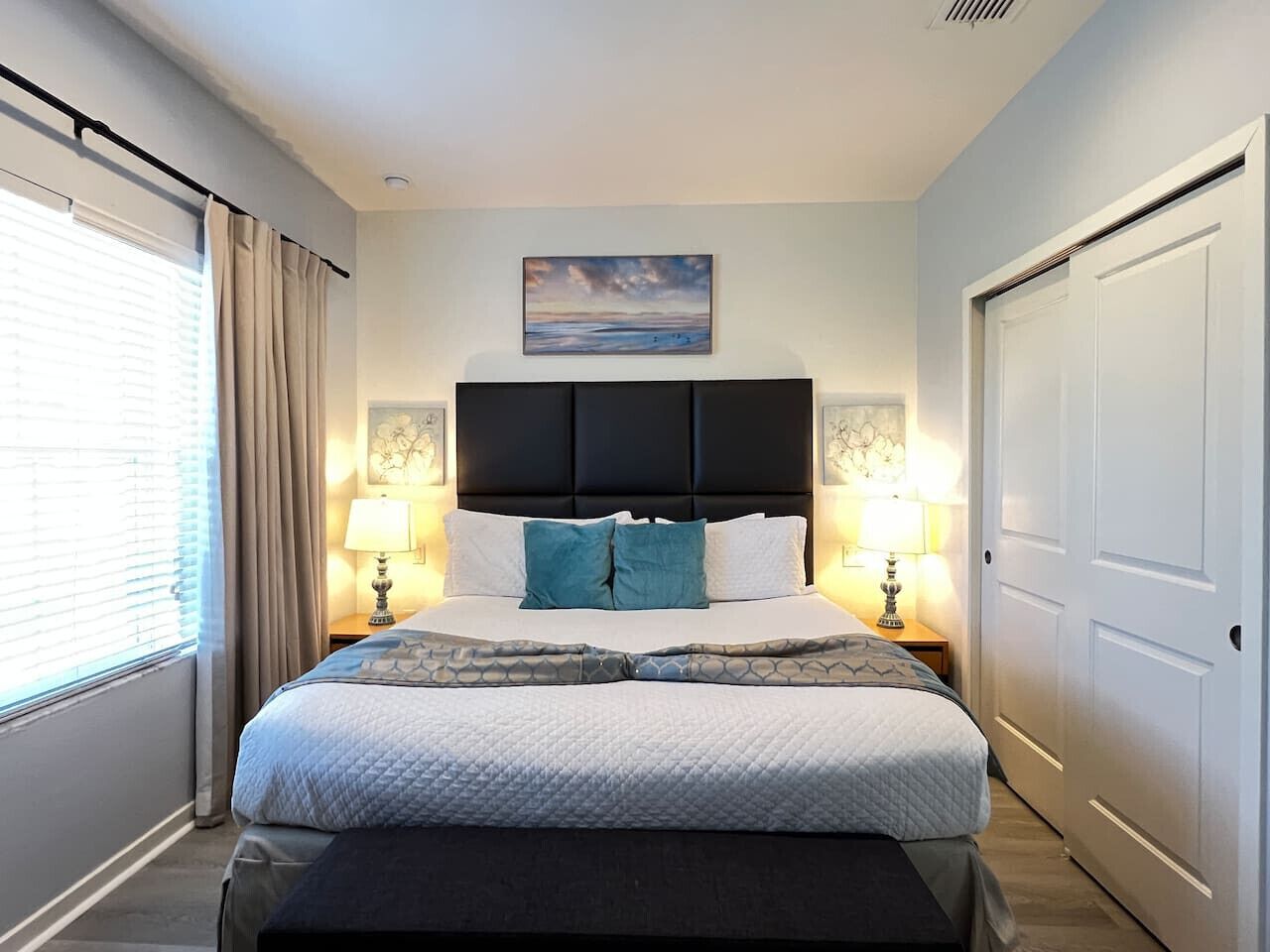 A neatly arranged bedroom with a bed centered between two bedside tables, each with a lamp. A painting hangs above the bed, and a door is visible on the right.