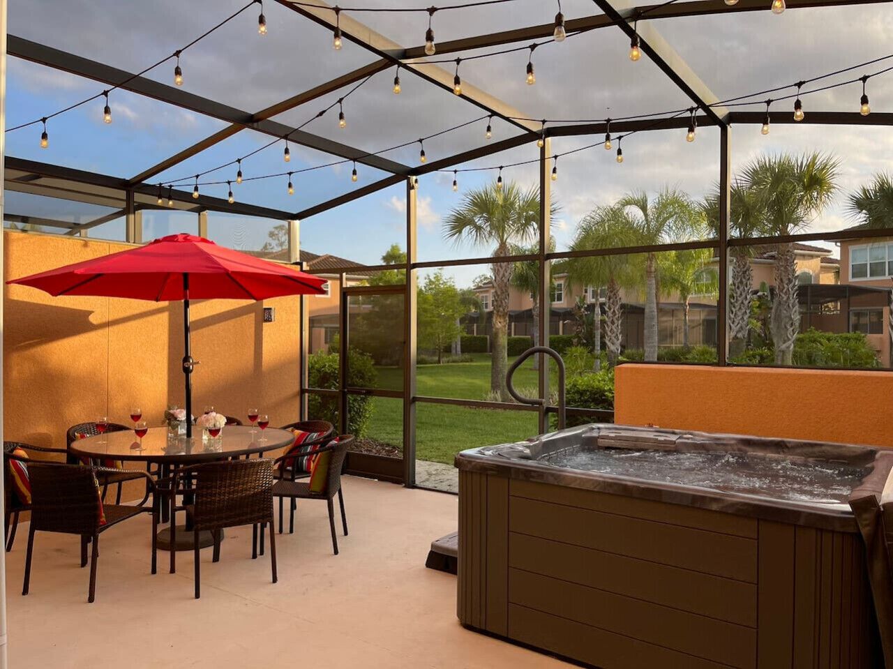 A covered outdoor patio with a hot tub, dining table, chairs, and a red umbrella.