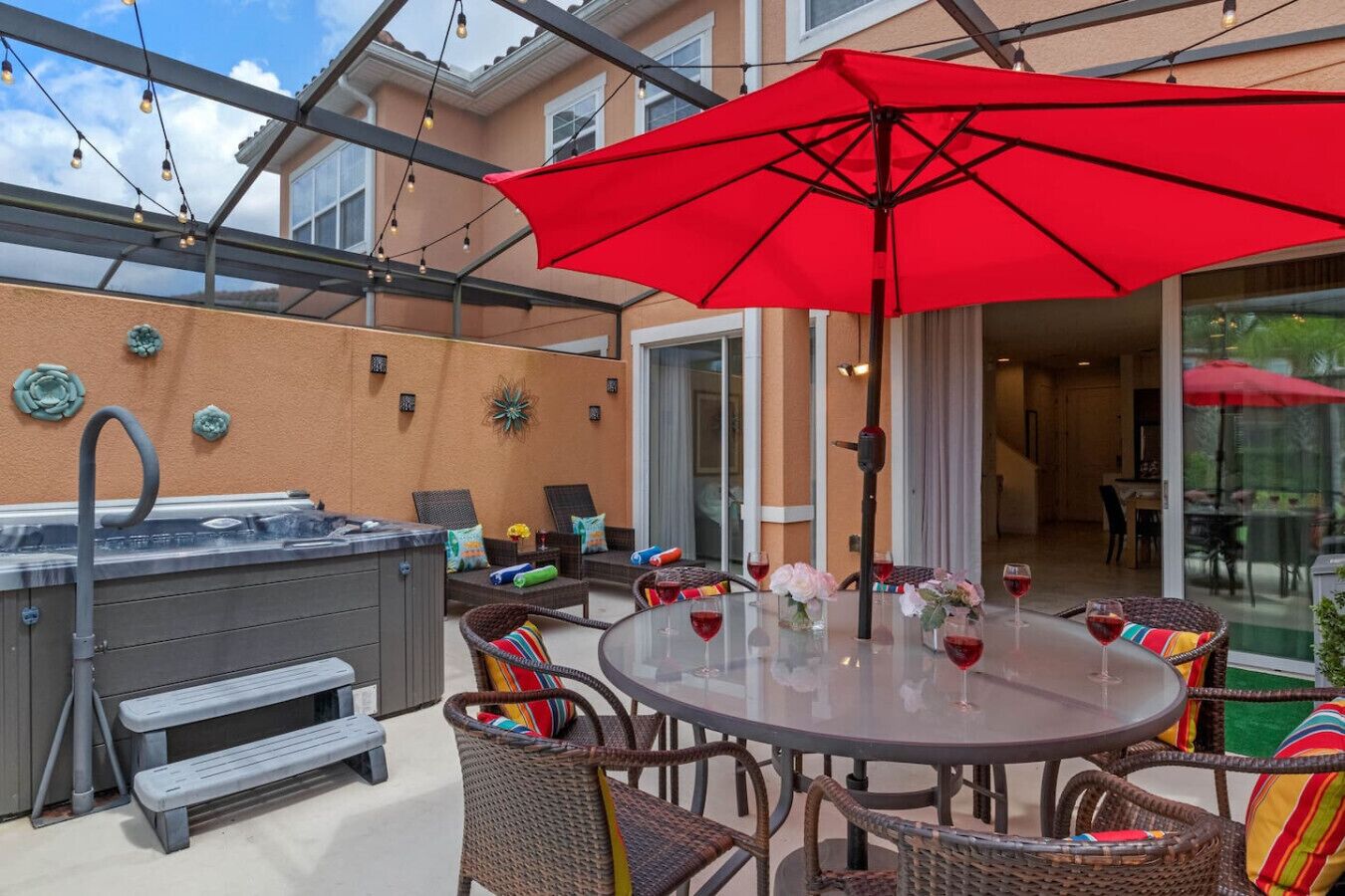 Un patio exterior con un jacuzzi, sillas de mimbre, una mesa redonda con copas de vino y un paraguas rojo.