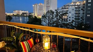 Een balkon met rieten meubilair, een tafel met een kaars, en uitzicht op een stadsbeeld met gebouwen en een waterpartij.