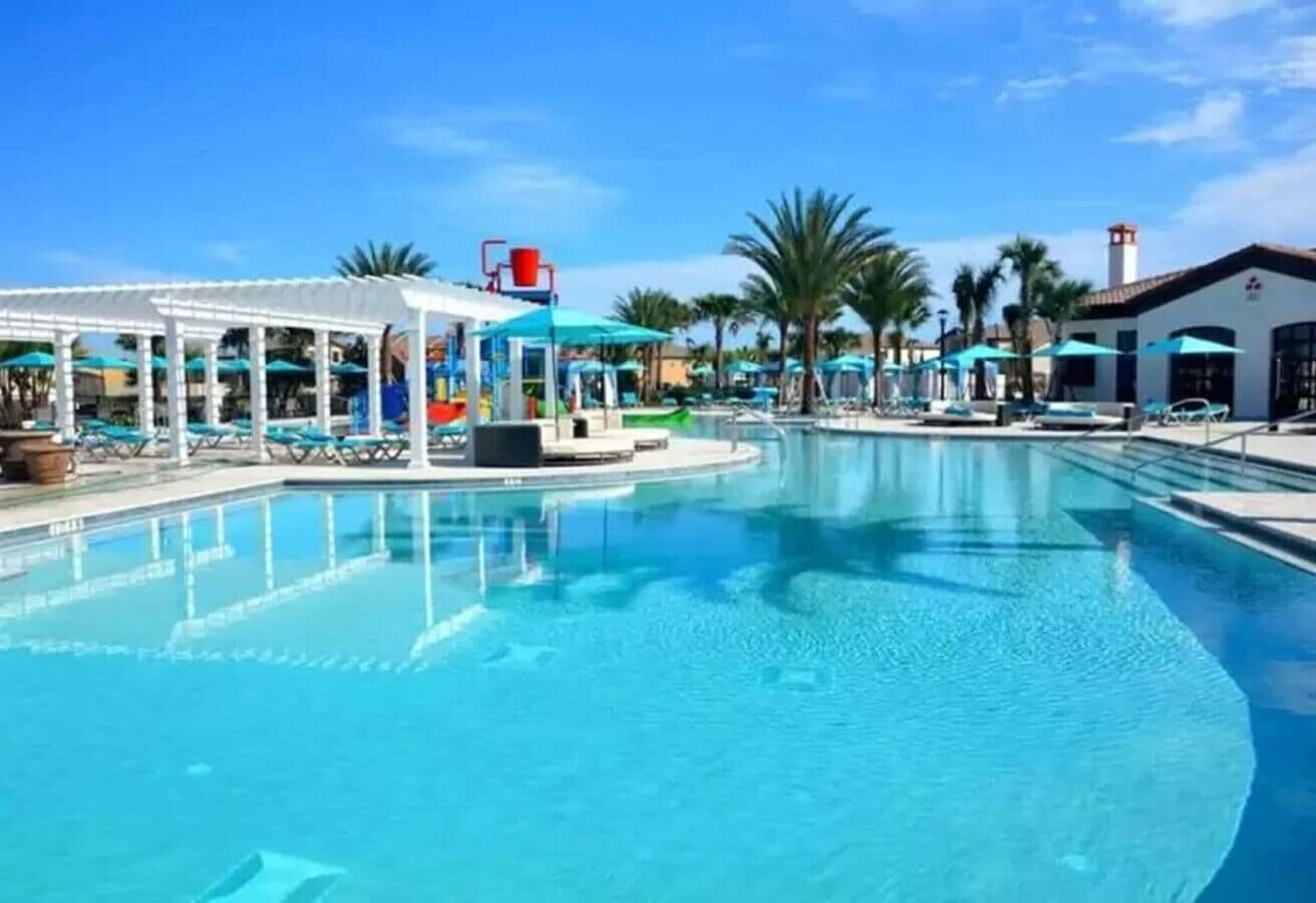 A large outdoor swimming pool with lounge chairs and umbrellas, surrounded by palm trees and a covered seating area.
