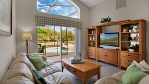 Uma sala de estar ampla com uma TV grande, móveis de madeira e vista para uma piscina e árvores.