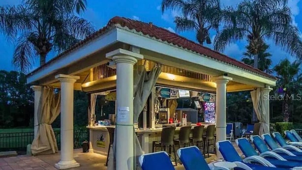 Uma área de estar coberta ao ar livre, com bar, poltronas azuis e palmeiras ao fundo.