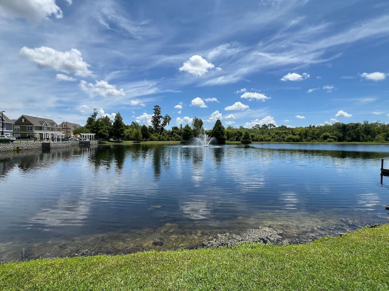 A calm lake with a fountain in the center, surrounded by a grassy area and trees.
