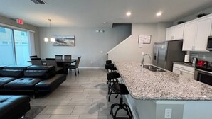 A modern kitchen with a granite countertop, stainless steel appliances, and a dining area with a black leather couch and chairs. - #5135 Cozy Villa 4/4 Centrally Located Disney/Epic (Kissimmee)