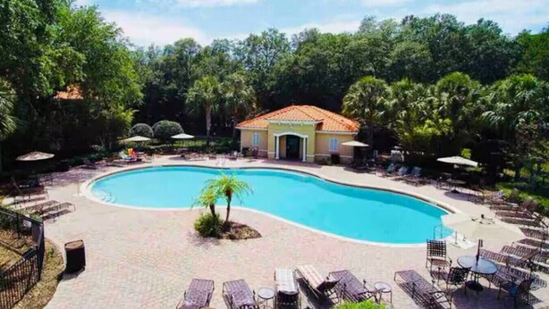 A swimming pool with lounge chairs and umbrellas, surrounded by trees and a building with a tiled roof. - #5135 Cozy Villa 4/4 Centrally Located Disney/Epic (Kissimmee)