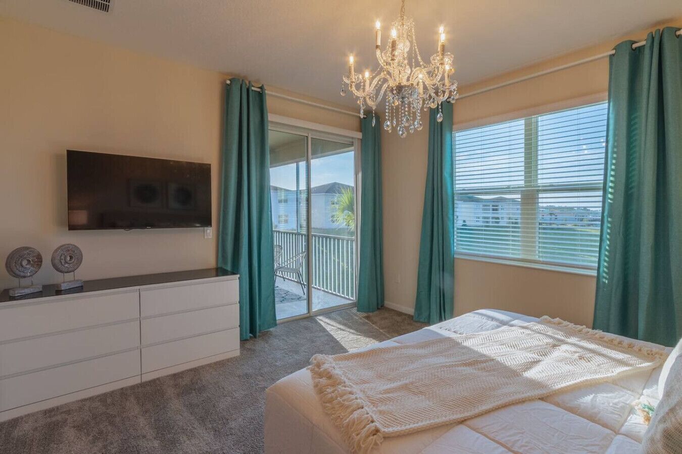 A bedroom with a large bed, a TV mounted on the wall, a dresser, and a balcony with green curtains.