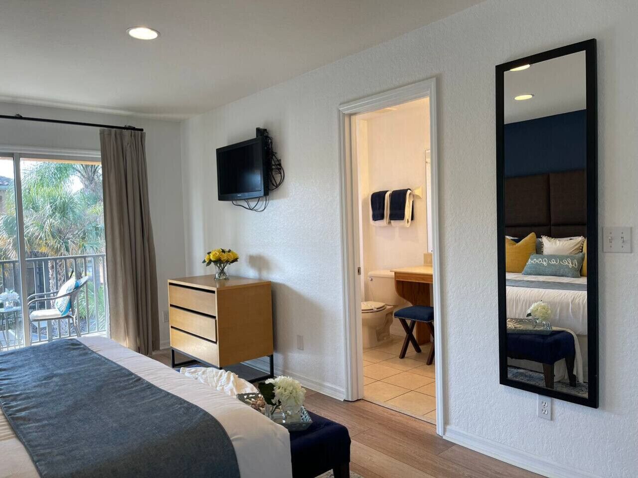 Ein Schlafzimmer mit einem großen Bett, einem Schminktisch, einem Fernseher und einem Spiegel. Es gibt einen Balkon mit einem Stuhl und einem Tisch.
