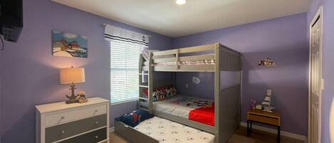 Una habitación con literas, un aparador, una lámpara, una ventana con persianas y una mesita con un juguete.