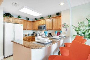 Une cuisine moderne avec des meubles en bois, un réfrigérateur en acier inoxydable, un four à micro-ondes et un comptoir en granit. On y trouve également un petit îlot central doté d’un évier et d’un tabouret.