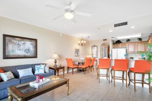 Un salon avec un canapé, une table basse et des fauteuils orange. On y trouve également un coin repas avec une table et des chaises, ainsi qu’une cuisine équipée de placards et d’un réfrigérateur.