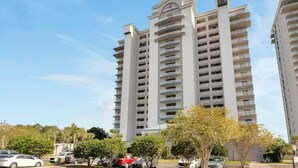 Un edificio residenziale a più piani con numerosi balconi, un parcheggio con auto e vegetazione circostante.