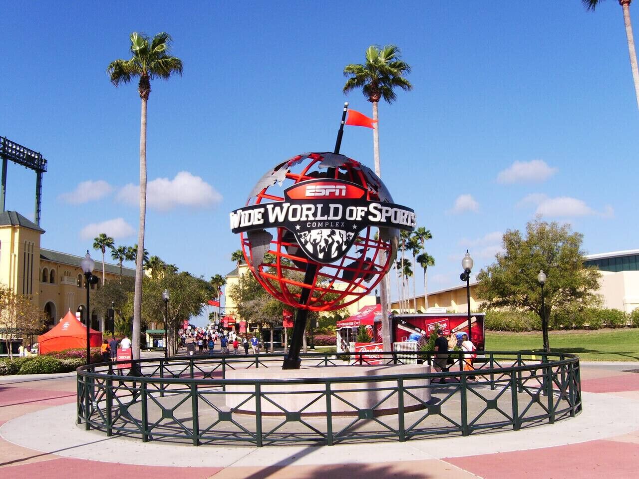 Eine große, kugelförmige Struktur mit dem Schriftzug "ESPN The World of Sports" und einer Flagge auf der Spitze, umgeben von einem grünen Zaun auf einem öffentlichen Platz mit Palmen und Gebäuden im Hintergrund.