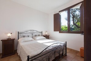 A bedroom with a metal bed frame, a bedside table with a lamp, a window with curtains, and a view of greenery outside. - Villa 'I Sogni Di Venere' with Sea View, Wi-Fi and Air Conditioning (Torre Suda)
