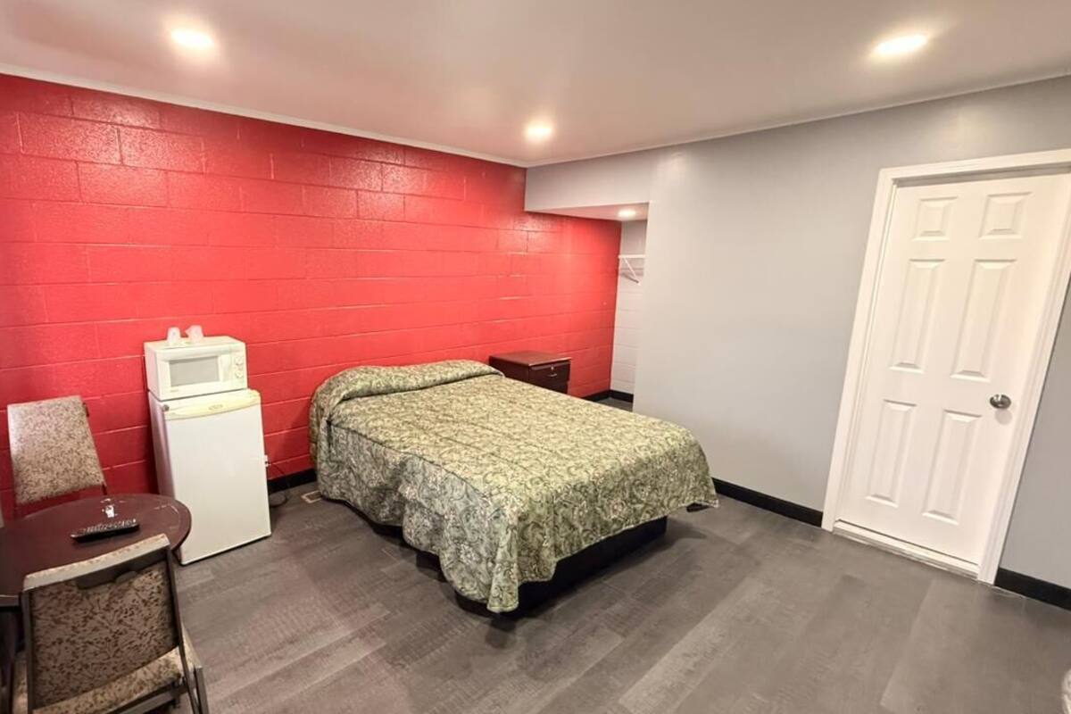 A small, single-bed room with a red brick wall, a white door, a small desk with a chair, and a bedside table.