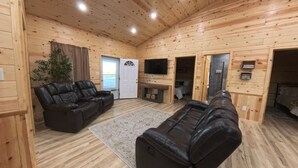 Ein Blockhaus-Interieur mit Kamin, Ledersofas, einem Flachbildfernseher und einer kleinen Küchenzeile.