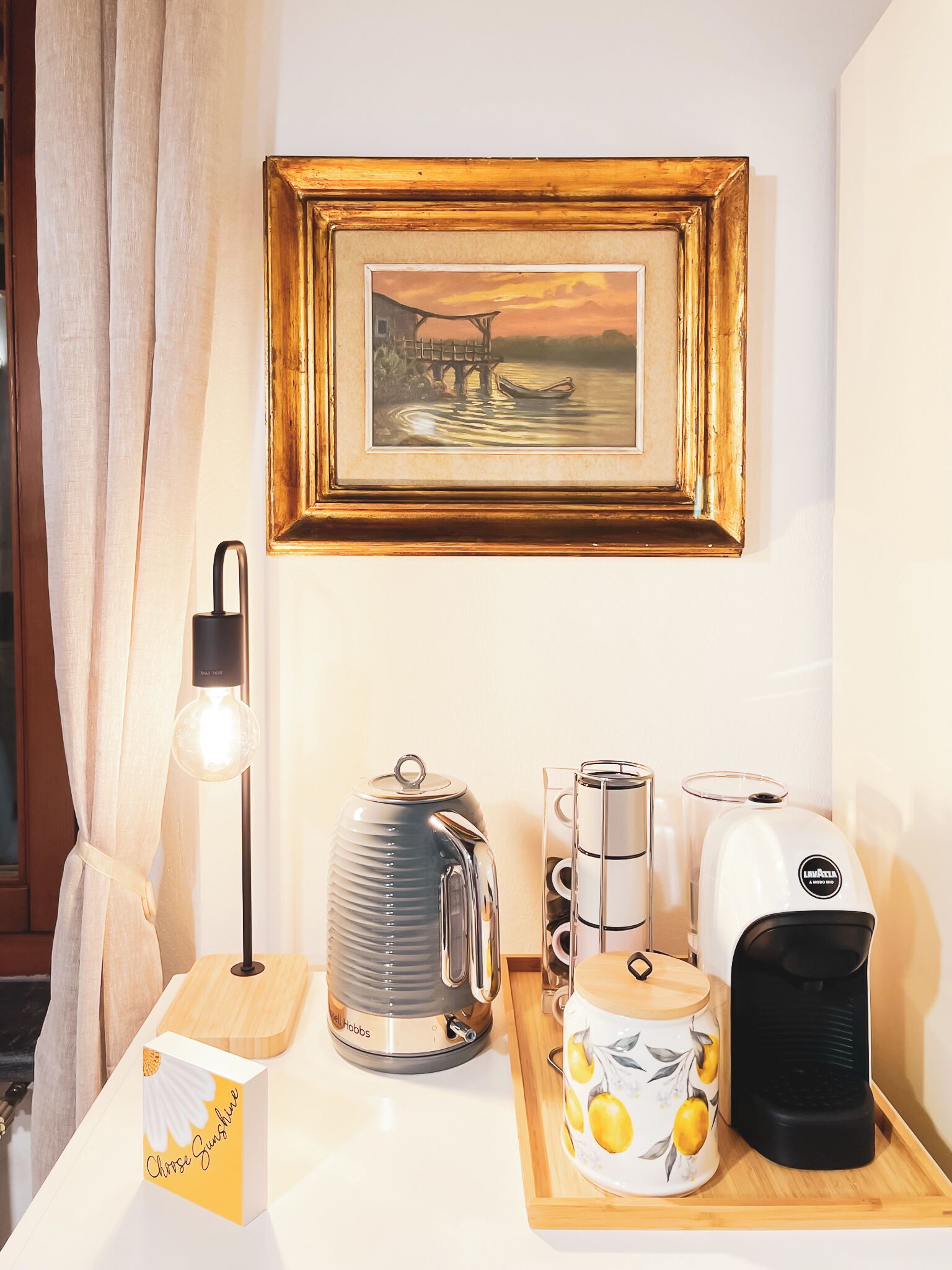 Un desayuno ordenado con hervidor, cafetera y un cuadro enmarcado sobre una mesa.
