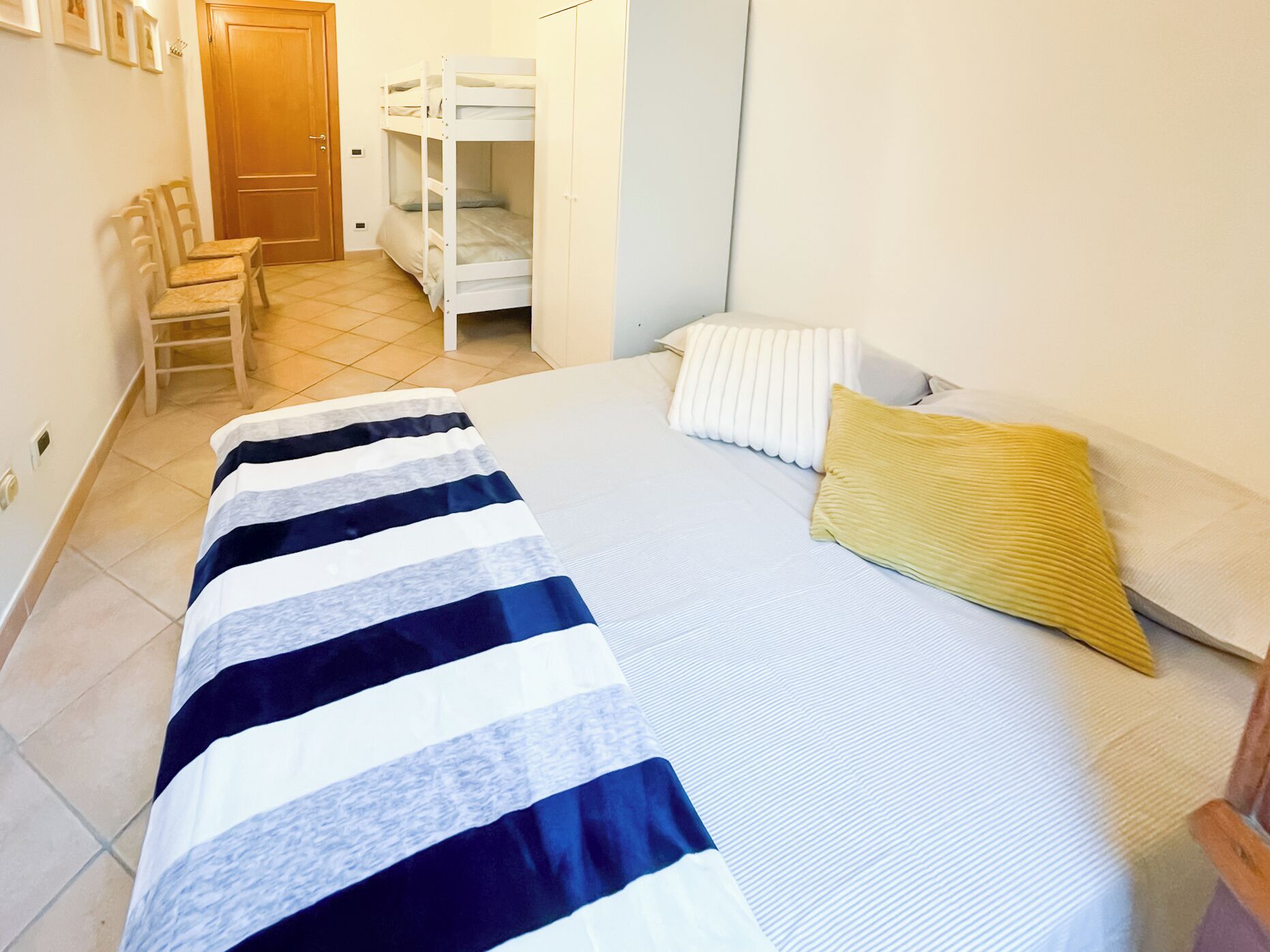 A single-bed room with a striped bedspread, a yellow pillow, and a white headboard. There is a wooden chair and a door in the background.