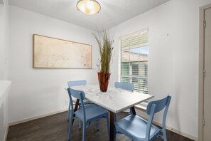 A dining area with a white table, blue chairs, a vase with plants, a painting on the wall, and a window with blinds. - Quiet Central Austin Getaway Near UT (Austin)
