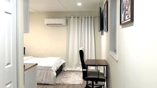 Habitación compacta con cama, escritorio, silla, aire acondicionado y ventana con cortinas.