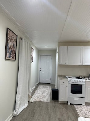 A narrow hallway with a white door at the end, a small kitchen area with a stove and cabinets, and a patterned rug on the floor.
