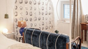 Ein Schlafzimmer mit einem Bett, einer blauen Sitzbank, einer Wand mit Muster, einer Chandelierleuchtre und einem Fenster mit Vorhängen.