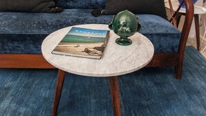 Ein blaues Sofa mit zwei dunklen Kissen, ein runder Tisch mit einem Buch und einer grünen Vase sowie eine Lampe auf einem Beistelltisch.