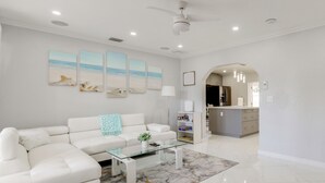 Un salon moderne avec un canapé blanc, une table basse en verre et un tapis à motifs de marbre. Trois tableaux sur le thème de la plage ornent le mur, et un ventilateur de plafond est installé au centre.